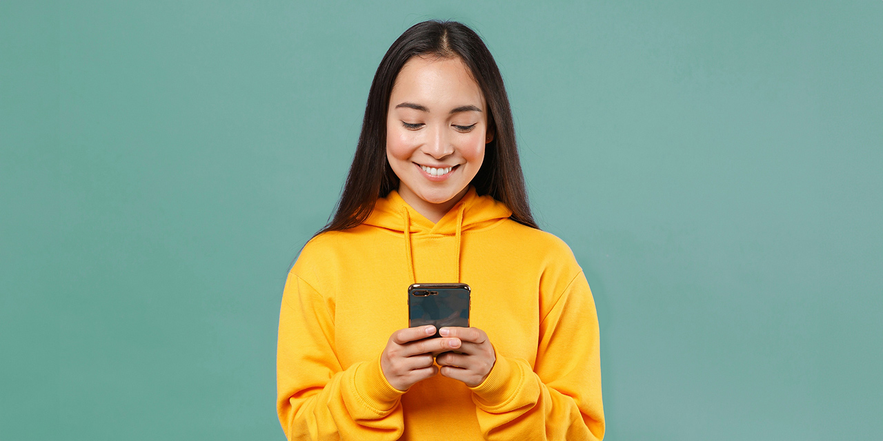 Junge Frau mit schwarzen langen Haaren und gelbem Kapuzenpulli sieht lächelnd in ihr Smartphone, das sie mit beiden Händen hält. - NettoKOM