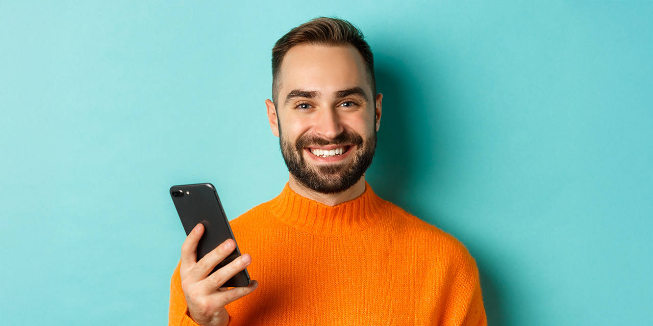 Junger Mann mit Bart hält lächelnd ein Smartphone vor hellblauem Hintergrund