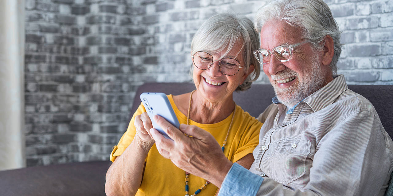 Älteres Paar sitzt auf dem Sofa, beide lächeln und schauen gemeinsam auf ein Smartphone.